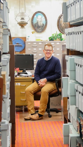 A smiling man in a blue sweater and tan chinos sits in front of a desk. He is looking down an aisle of blue boxes containing archival documents. Behind him is a painted portrait of a man with a white beard as well as other historical artifacts.